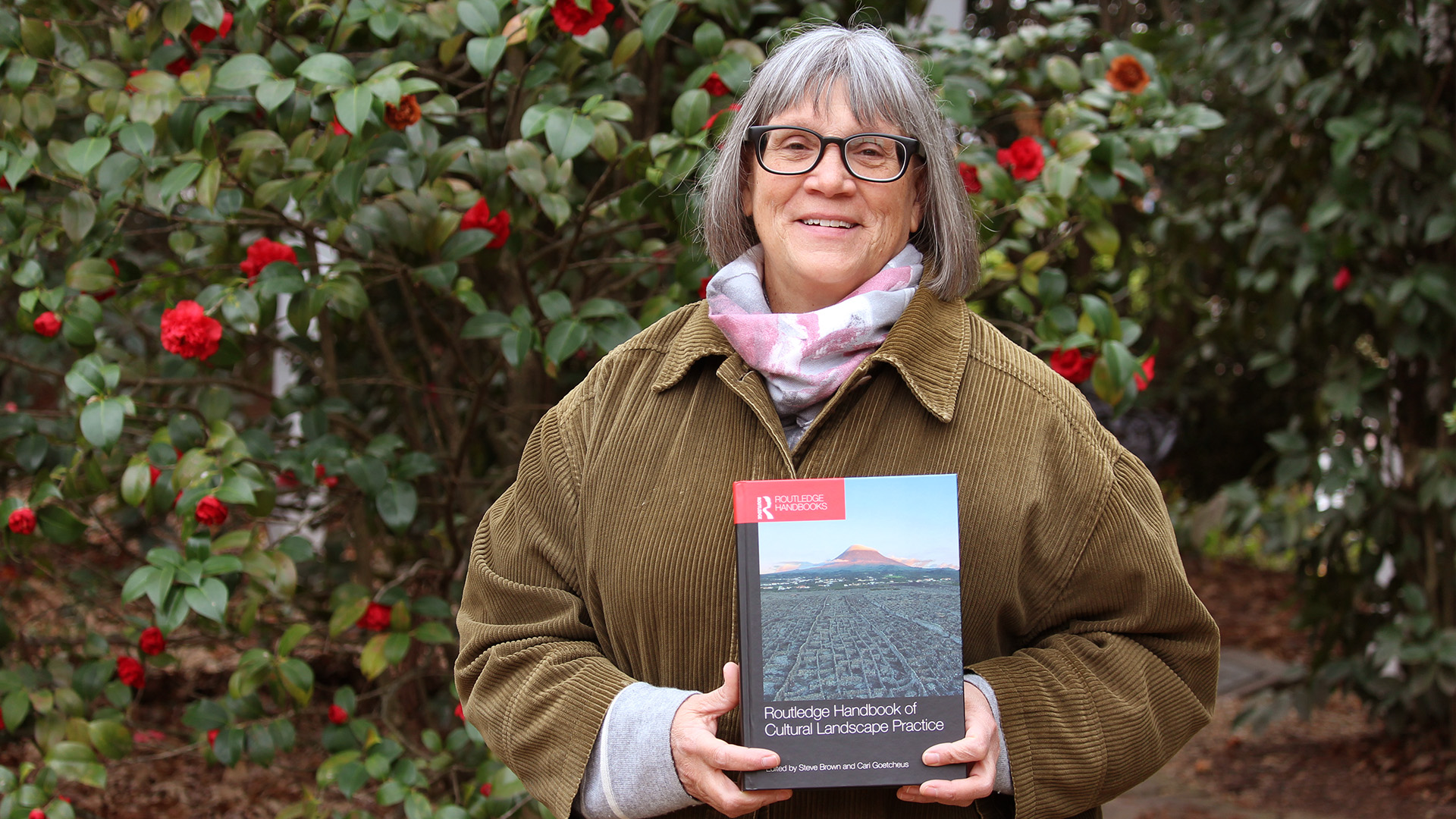 CED professor Cari Goetcheus stands before a camellia bush holding "The Routledge Handbook of Cultural Landscape Practice." 
