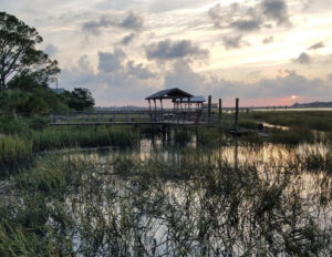 Wooden dock with covered seating extends over a coastal marsh at sunset, surrounded by tall grasses and calm tidal water.