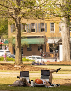 Two students lying on the campus lawn with laptops, studying under trees near downtown buildings.