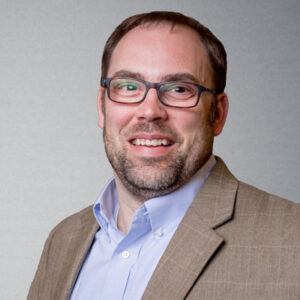 Portrait of Buck Bacon, Government and Community Services Director at W&A Engineering, wearing glasses, a light blue shirt, and a tan blazer.