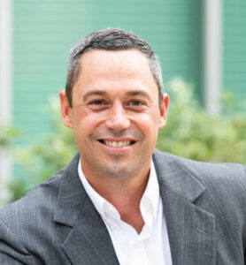 Russ Seamon wearing a gray suit and white shirt smiles while posing outdoors with greenery in the background.