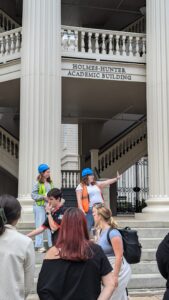 Students wearing safety vests and blue hard hats stand on the steps of the Holmes-Hunter Academic Building at UGA, speaking to a group during a campus experience tour.