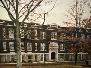 A three-story, dark brick and stone building, a historic school, is centered in this sepia-toned photograph, viewed through a row of bare trees. A prominent white stone arched entryway with ornate details is visible in the middle of the ground floor.